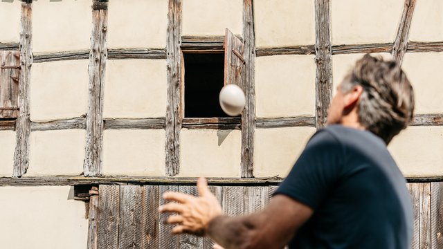Ein Mann wirft einen Ball vor einem schönen Fachwerkhaus. Neue Höfe schafft regionale Werte neu.