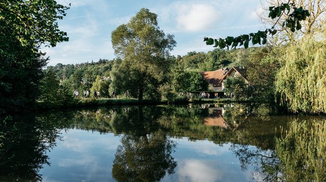 Landschaftsansicht: ein See von Bäumen umrandet, am hinteren Seerand sieht man ein Dorf. Menschen spazieren am Seeufer – Neuhof a. d. Zenn – fränkische Toskana und ländliche Idylle – Neue Höfe 